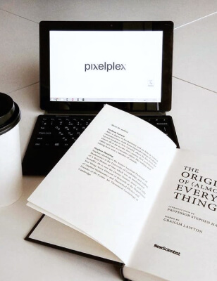 Нетбук с логотипом PixelPlex на столе рядом с книгой New Scientist: The Origin of (almost) Everything